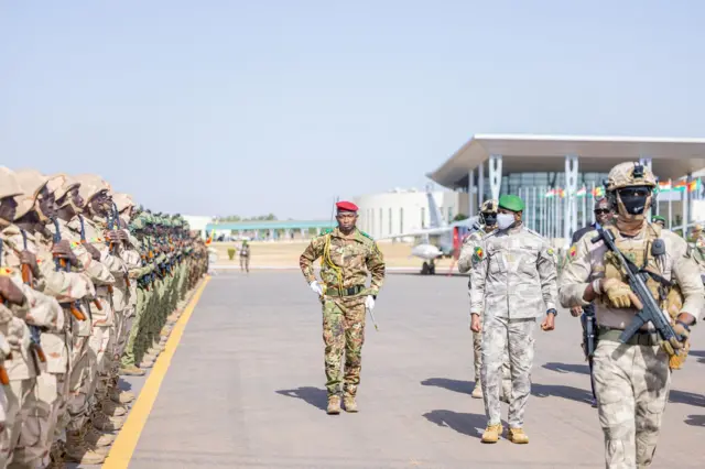 Le général Assimi Goïta, président de la Transition au Mali, passe en revue des troupes en tenue militaire avec un béret vert et un cache-nez.